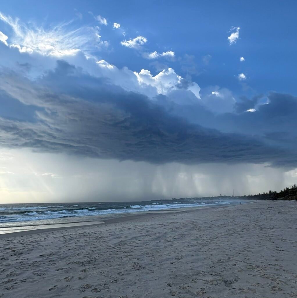 A dramatic storm cloud stretches over a quiet Gold Coast beach, with soft light breaking through and waves rolling gently to shore. The scene symbolises the emotional weight and shifting nature of complex mental health presentations.