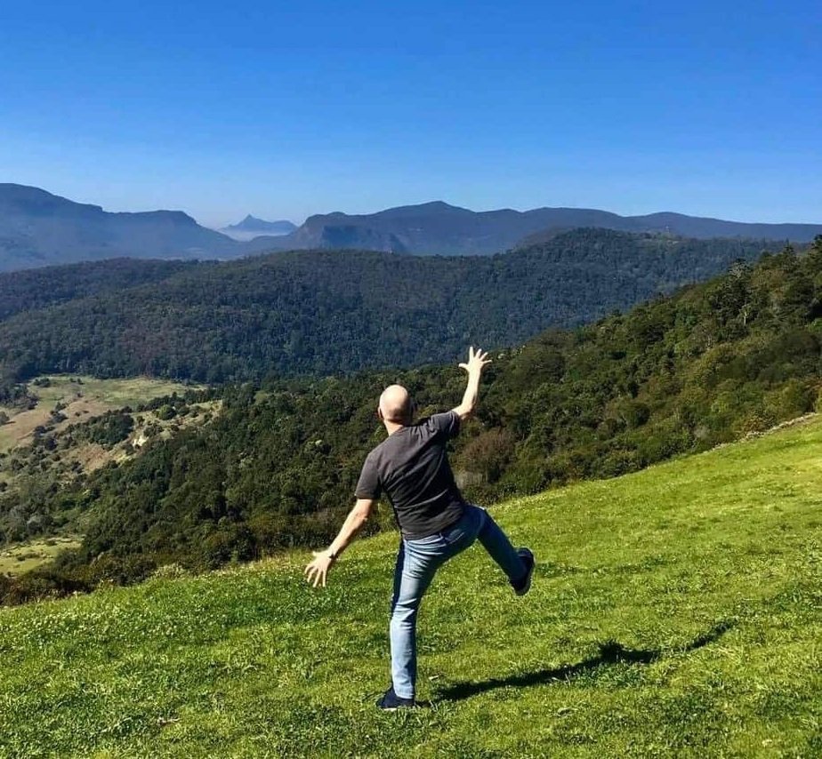 Psychology of fun | David the Psychologist stands playfully on a sunny hillside with one leg lifted and arms raised, overlooking a wide mountain landscape. The scene reflects a joyful moment in nature that aligns with the article about light hearted play and emotional wellbeing.