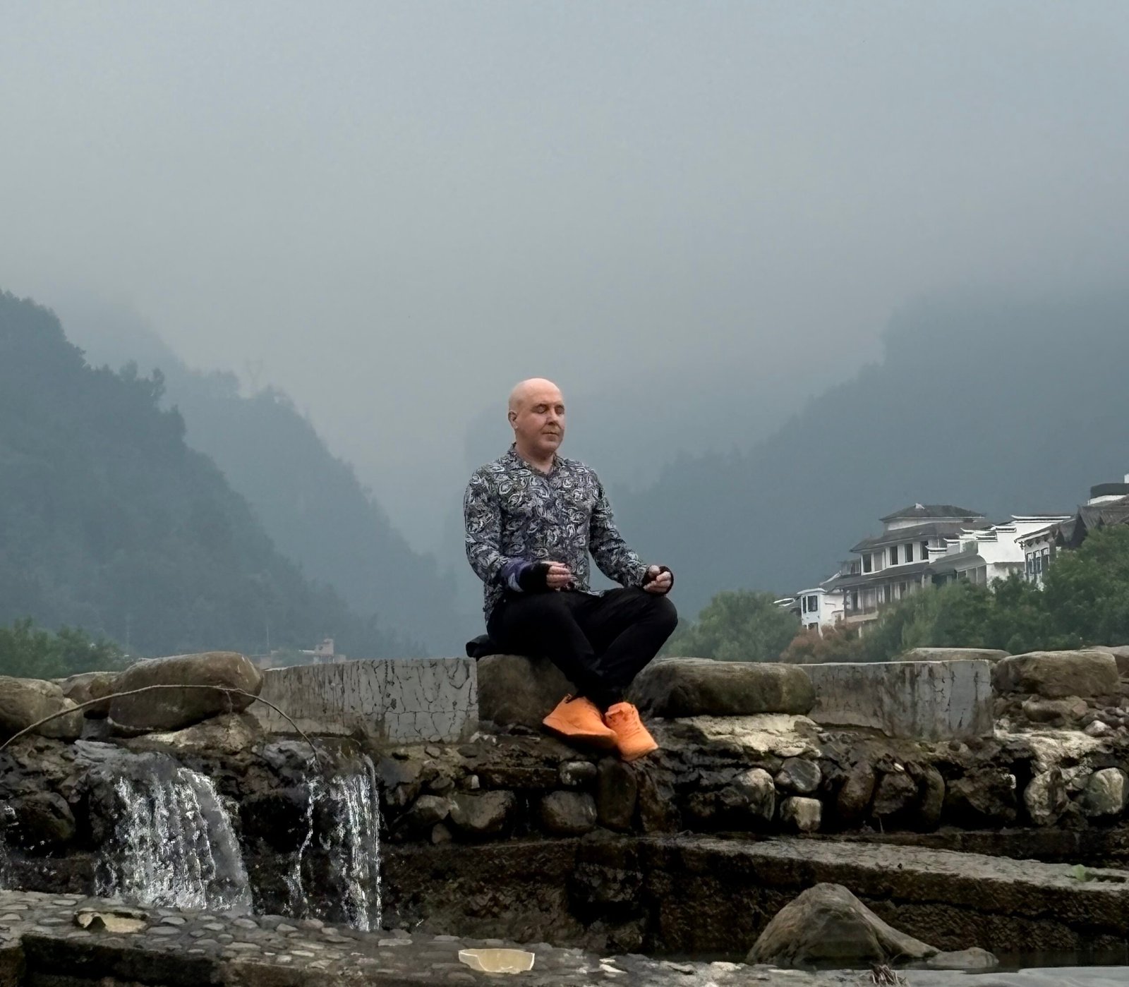 David the Psychologist sits peacefully by a small waterfall in a mountain village, wearing a colourful paisley shirt and orange shoes, practising mindfulness with a calm expression.