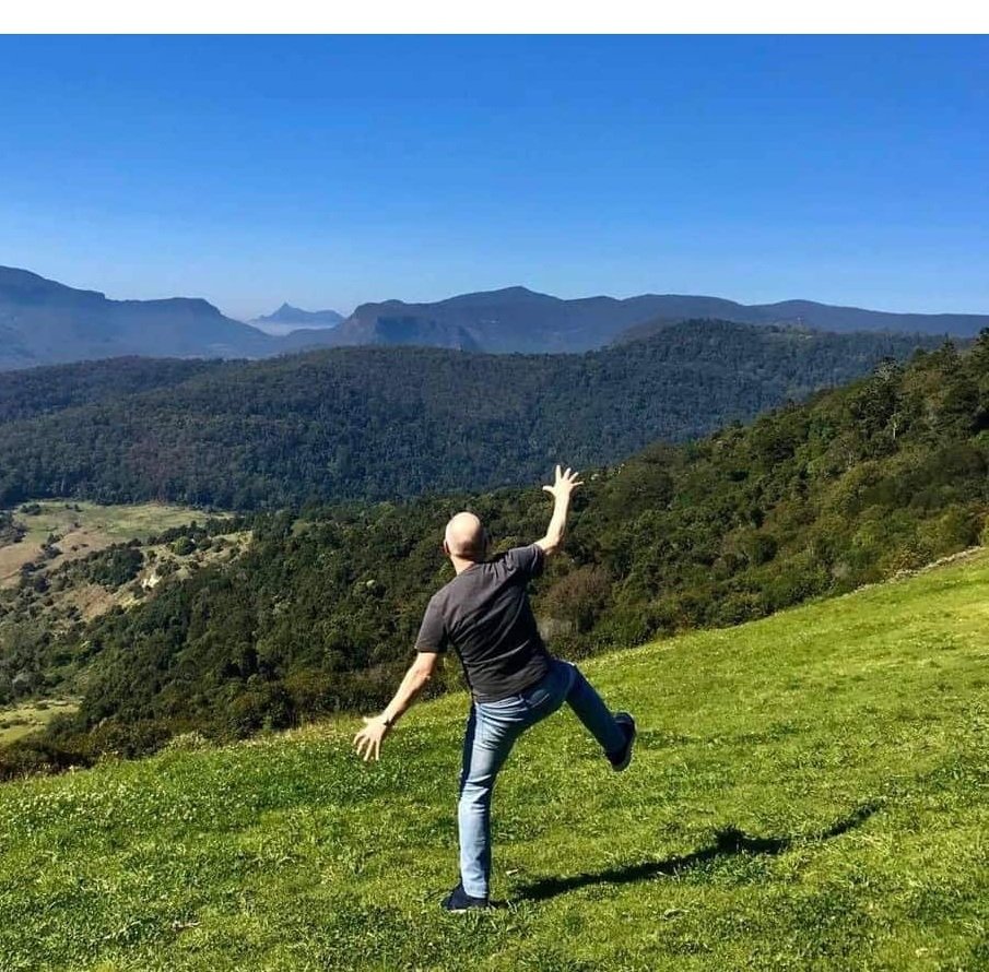 David the psychologist oyfully balancing and moving playfully on a grassy hillside overlooking mountains and forest, illustrating how fun, movement, and connection with nature support psychological well-being.