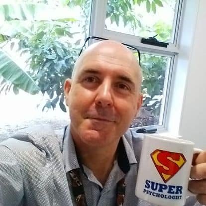 Get to Know David | David the Psychologist holding a white mug with a red and yellow S symbol and the words Super Psychologist, sitting in his Varsity Lakes office with trees visible through the window.