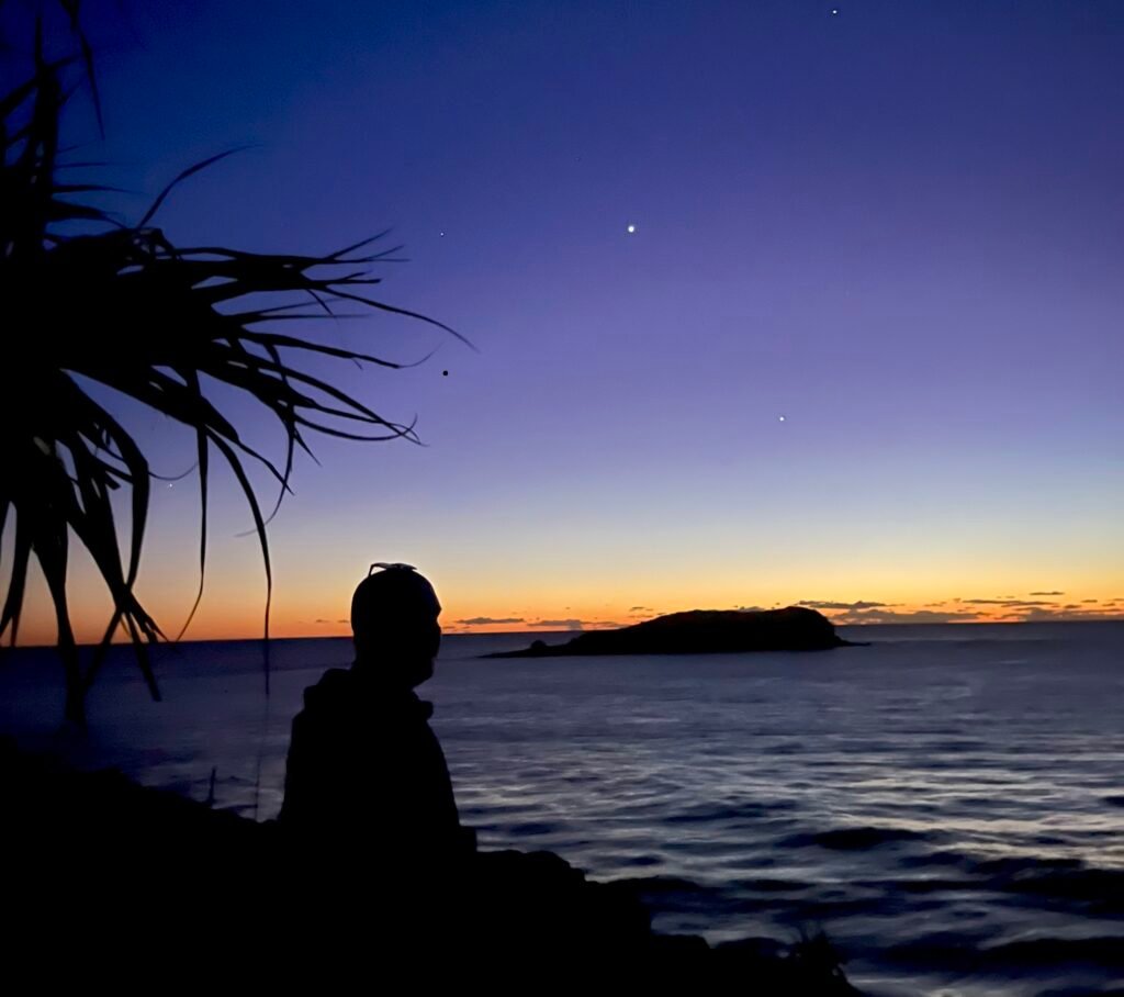 Therapist self-care | Man sitting at the oceanside with a bright orange sunrise beside him 