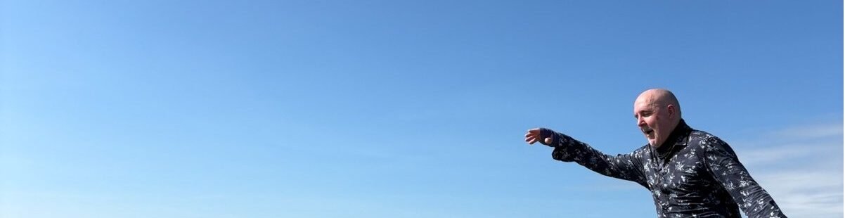 David the Psychologist balancing on a mountain peak under a clear blue sky, symbolising mindfulness, perspective, and balance when responding to the inner critic.