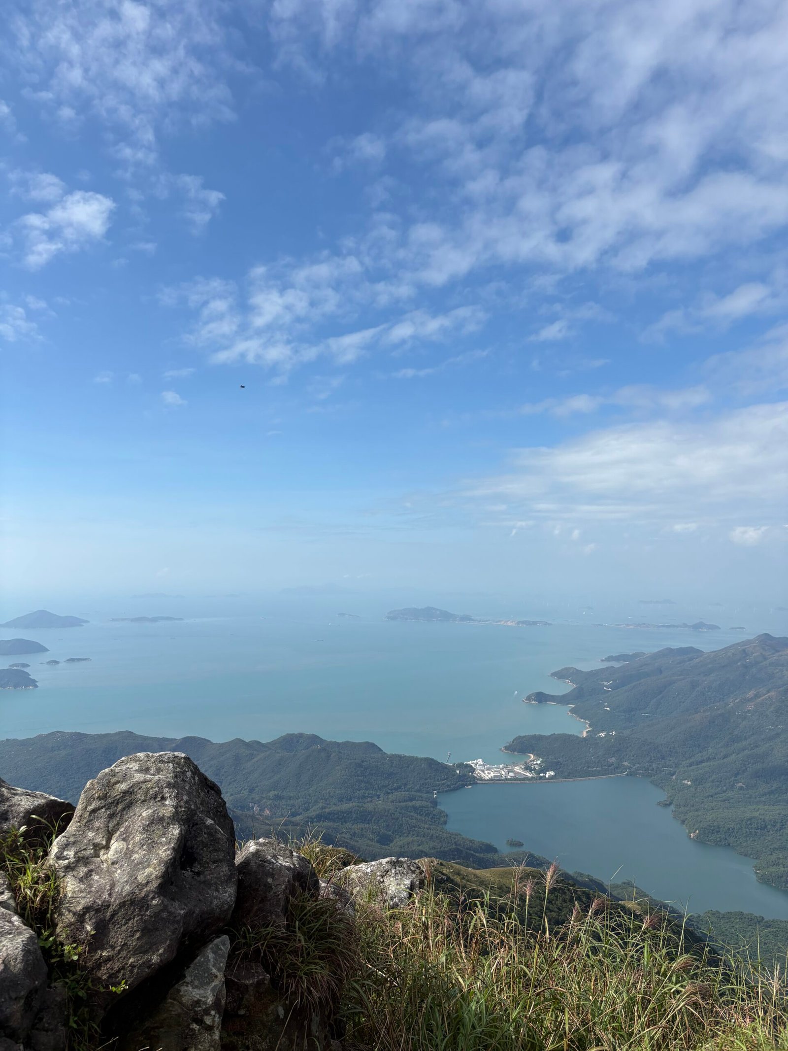A wide landscape view from a mountain peak showing blue sky, scattered clouds, and a calm ocean with distant islands, reflecting a sense of balance and spaciousness.
