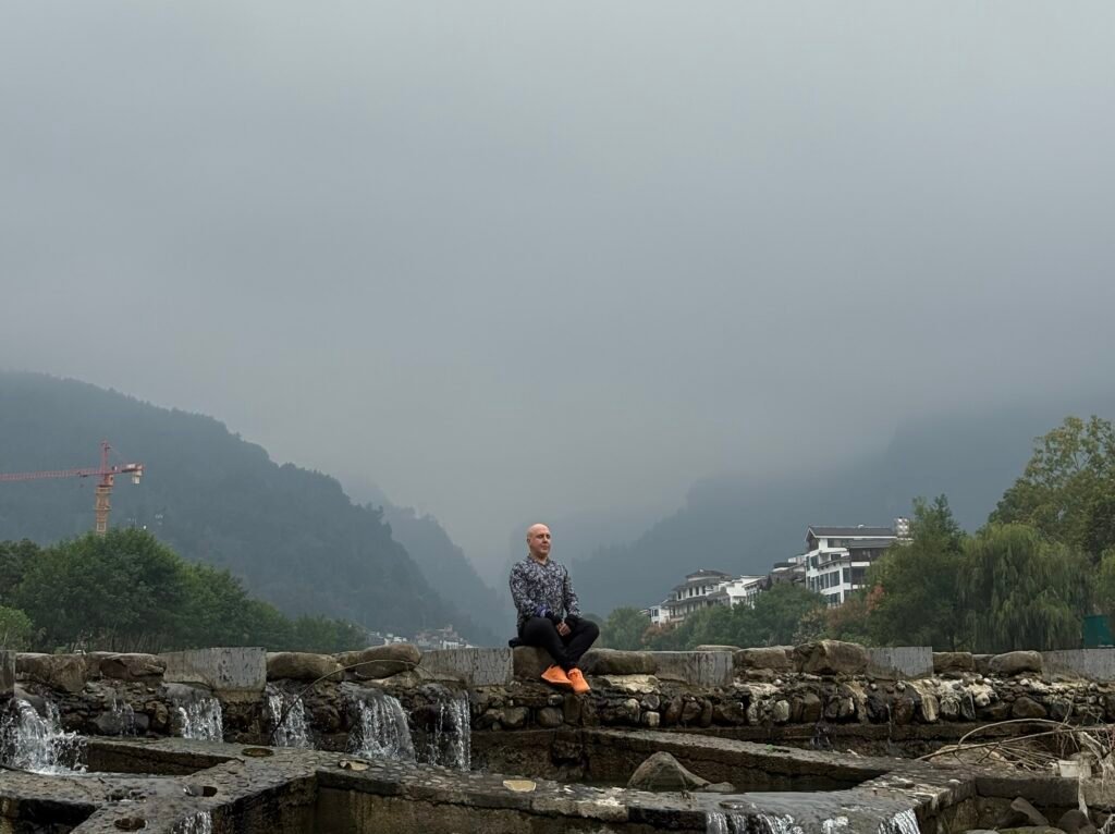 The Irony of Attachment | David the Psychologist sits calmly beside flowing water and misty mountains, representing the irony of attachment — seeking stability while learning to release grasping through awareness and balance.
