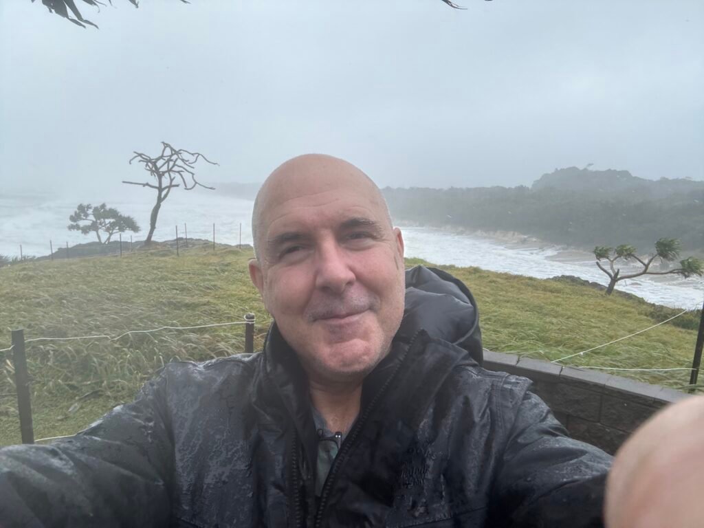 David the Psychologist smiling in a rainstorm near the coast, wearing a wet jacket, with windblown trees and rough ocean in the background.