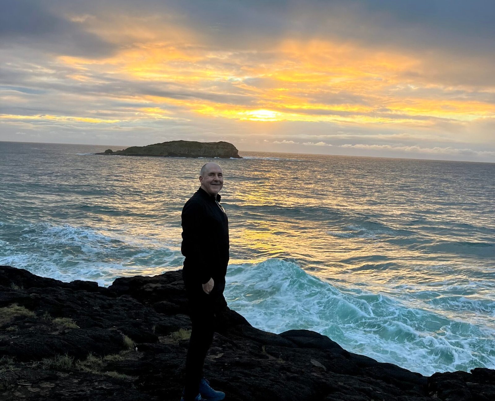David the psychologist standing on rocky coastline at sunrise, looking out over the ocean, representing reflection and recovery from burnout in helping professions.