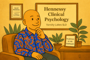 David Hennessy, Clinical Psychologist in Varsity Lakes, Gold Coast, seated in a therapy room wearing a colourful paisley shirt, with Hennessy Clinical Psychology signage visible, offering therapy for social anxiety in person or via telehealth.